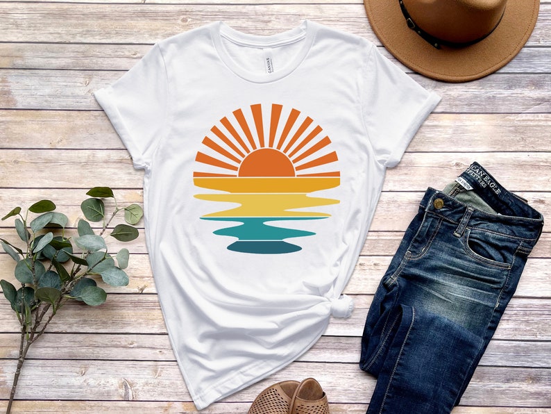May include: A white t-shirt with a retro sunset graphic featuring a sun with rays and a reflection in the water. The graphic is in shades of orange, yellow, blue, and green.