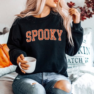 May include: A black sweatshirt with the word "SPOOKY" printed in orange on the front. The sweatshirt is being worn by a person with long blonde hair. The person is sitting on a white couch with a pillow behind them.