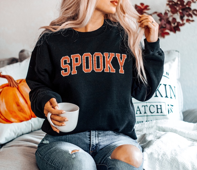 May include: A black sweatshirt with the word "SPOOKY" printed in orange on the front. The sweatshirt is being worn by a person with long blonde hair. The person is sitting on a white couch with a pillow behind them.
