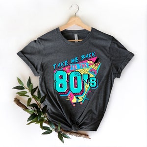 Op de afbeelding: Een donkergrijs T-shirt met een grafische print die "Take Me Back To The 80's" zegt in een retro-stijl met kleurrijke geometrische vormen en een cassettebandje.
