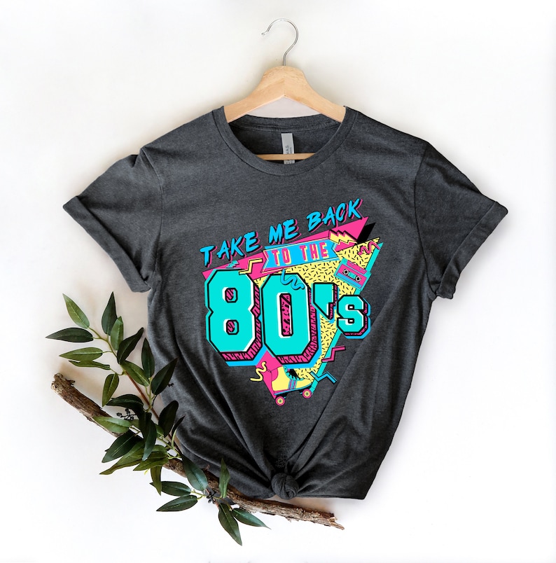 Op de afbeelding: Een donkergrijs T-shirt met een grafische print die "Take Me Back To The 80's" zegt in een retro-stijl met kleurrijke geometrische vormen en een cassettebandje.