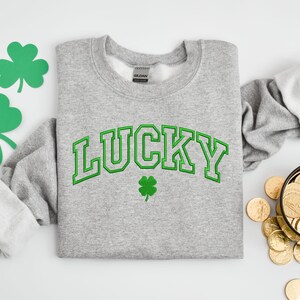 K&ouml;nnte beinhalten: Ein grauer Pullover mit dem Wort "LUCKY" in gr&uuml;nen Buchstaben und einem gr&uuml;nen Kleeblatt.
