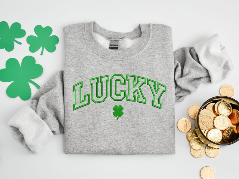 K&ouml;nnte beinhalten: Ein grauer Pullover mit dem Wort "LUCKY" in gr&uuml;nen Buchstaben und einem gr&uuml;nen Kleeblatt.