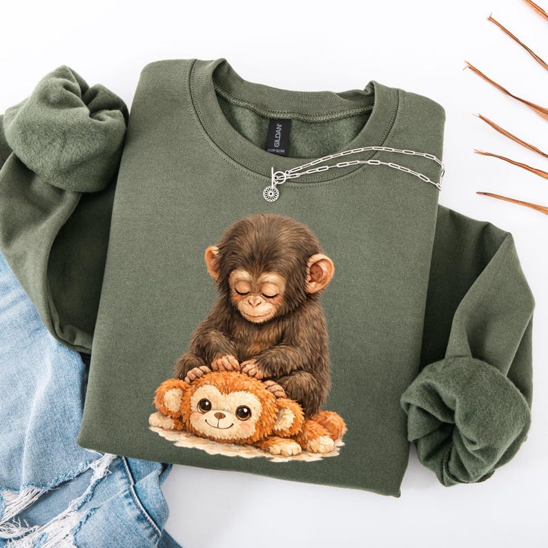 May include: Olive green sweatshirt featuring a detailed illustration of a brown monkey with closed eyes, gently touching a smaller, orange monkey. A silver necklace with a circular pendant rests on the sweatshirt. The sweatshirt is folded on a white surface.