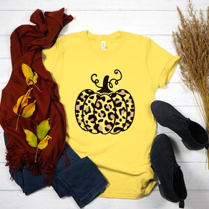 May include: A yellow t-shirt with a black and brown leopard print pumpkin graphic. The pumpkin has a stem and curly vines.