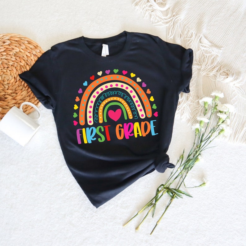 Peut inclure: T-shirt noir avec un motif arc-en-ciel et le texte "FIRST GRADE" en lettres color&eacute;es. L'arc-en-ciel a un c&oelig;ur au centre.