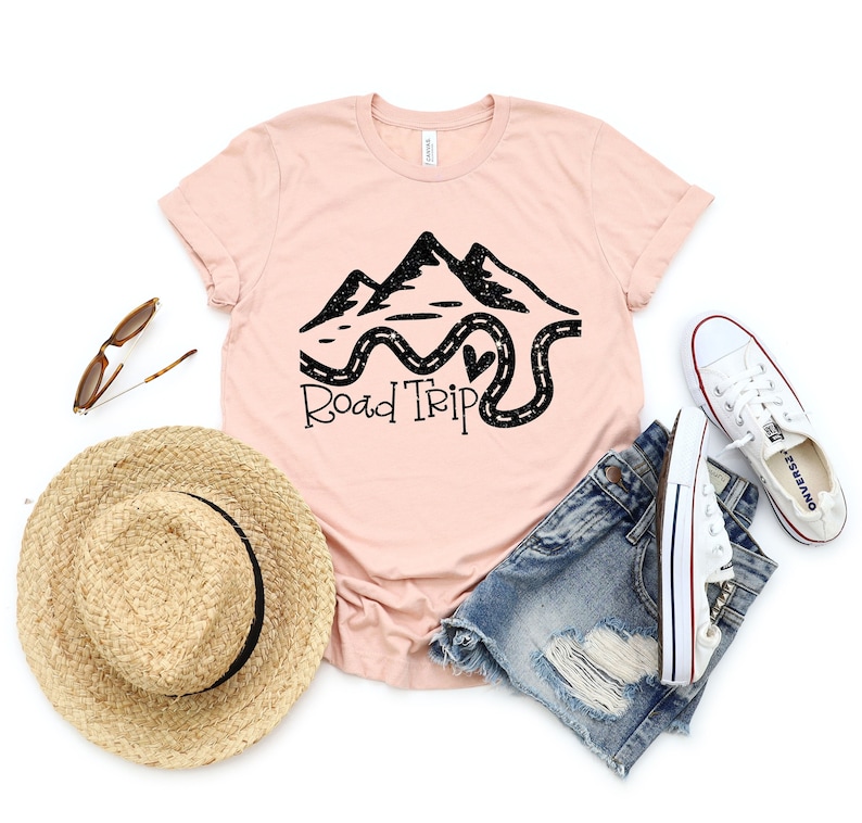 Puede incluir: Una camiseta rosa con un gr&aacute;fico negro de monta&ntilde;as y un camino sinuoso con el texto "Road Trip".