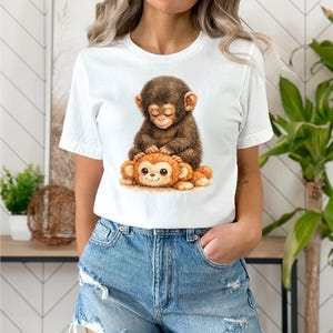 May include: White t-shirt featuring a watercolor illustration of two monkeys. One brown monkey is sitting on top of a smaller, orange monkey. The design has a cute, whimsical style, perfect for animal lovers.