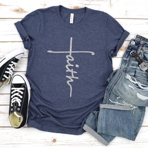 May include: A navy blue t-shirt with a white cross and the word "faith" written vertically on the cross. The shirt is laid flat on a white wooden surface with a pair of black sneakers and a pair of blue jeans.