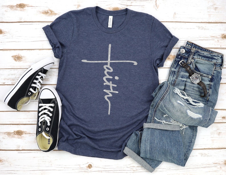 May include: A navy blue t-shirt with a white cross and the word "faith" written vertically on the cross. The shirt is laid flat on a white wooden surface with a pair of black sneakers and a pair of blue jeans.