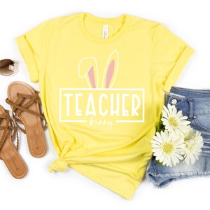 May include: A yellow t-shirt with a white graphic that says "Teacher bunny" with pink bunny ears above the text. The shirt is laid out on a white background with a pair of brown sandals, sunglasses, and denim shorts with white flowers.