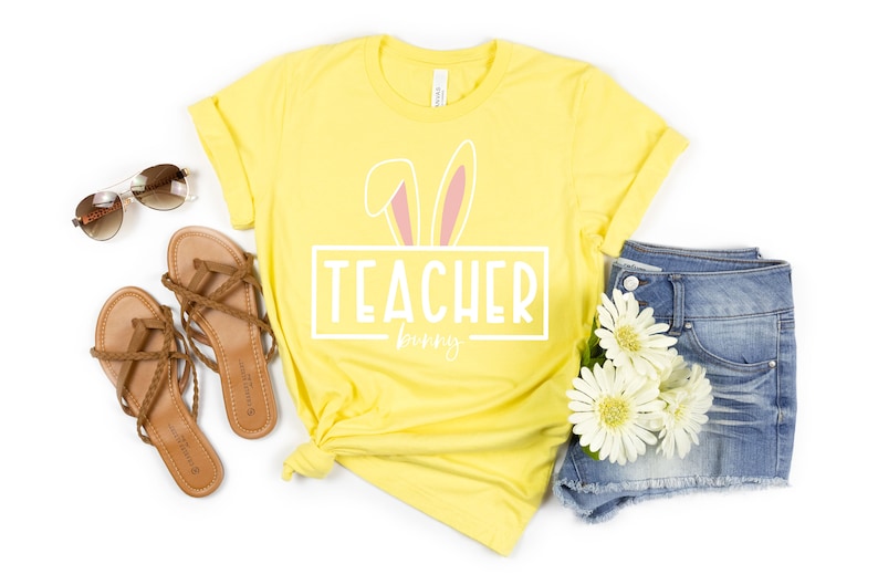May include: A yellow t-shirt with a white graphic that says "Teacher bunny" with pink bunny ears above the text. The shirt is laid out on a white background with a pair of brown sandals, sunglasses, and denim shorts with white flowers.