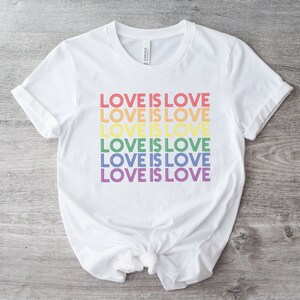 Może przedstawiać: Biała koszulka z napisem "LOVE IS LOVE" powt&oacute;rzonym sześć razy w kolorach tęczy.