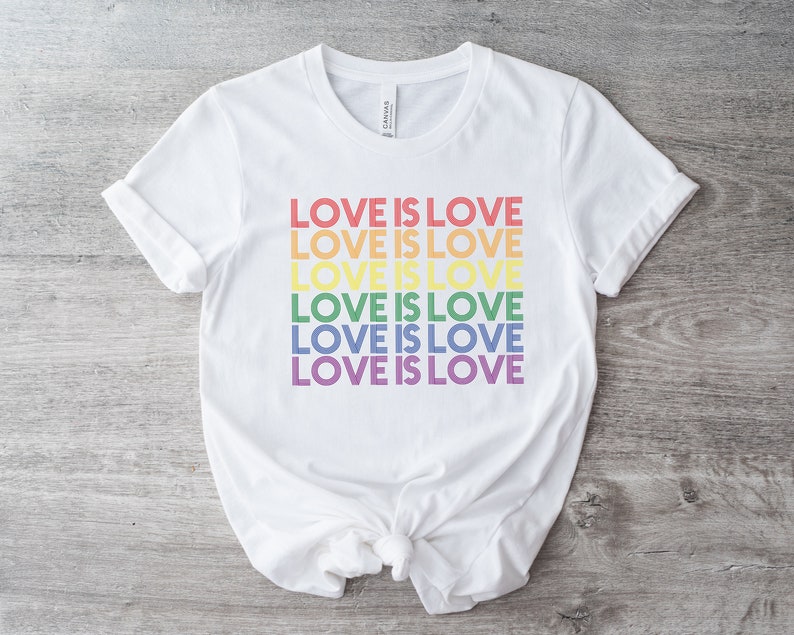Może przedstawiać: Biała koszulka z napisem "LOVE IS LOVE" powt&oacute;rzonym sześć razy w kolorach tęczy.