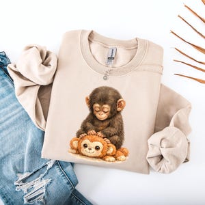 May include: A beige sweatshirt featuring a watercolor illustration of a baby monkey hugging a stuffed monkey. The design is in shades of brown and orange. The sweatshirt is paired with blue denim jeans.
