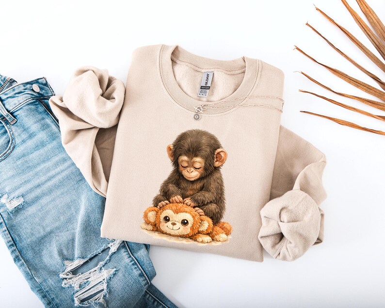 May include: A beige sweatshirt featuring a watercolor illustration of a baby monkey hugging a stuffed monkey. The design is in shades of brown and orange. The sweatshirt is paired with blue denim jeans.