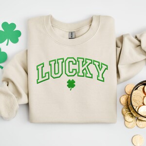 K&ouml;nnte beinhalten: Ein cremefarbener Pullover mit dem Wort "LUCKY" in gr&uuml;nen umrandeten Buchstaben. Unter dem Wort befindet sich ein kleines gr&uuml;nes Kleeblatt.