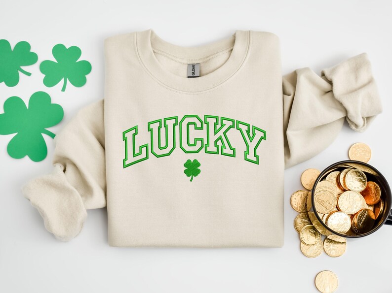 K&ouml;nnte beinhalten: Ein cremefarbener Pullover mit dem Wort "LUCKY" in gr&uuml;nen umrandeten Buchstaben. Unter dem Wort befindet sich ein kleines gr&uuml;nes Kleeblatt.