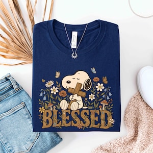 Puede incluir: Camiseta azul marino con un dise&ntilde;o de dibujos animados de Snoopy sosteniendo una cruz, rodeado de flores y mariposas. La palabra "BLESSED" est&aacute; impresa en marr&oacute;n con una fuente retro. Se ve un collar plateado con un colgante de sol.