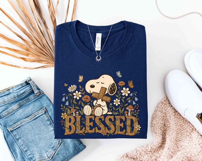 Puede incluir: Camiseta azul marino con un dise&ntilde;o de dibujos animados de Snoopy sosteniendo una cruz, rodeado de flores y mariposas. La palabra "BLESSED" est&aacute; impresa en marr&oacute;n con una fuente retro. Se ve un collar plateado con un colgante de sol.