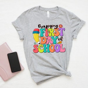 May include: A gray t-shirt with the text "happy first day of school" in colorful letters. A cartoon pencil with a red eraser is behind the text.