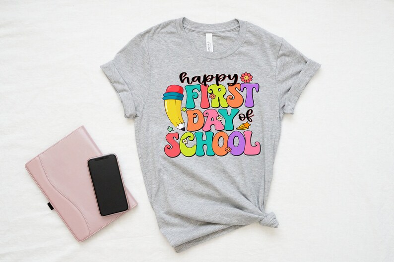 May include: A gray t-shirt with the text "happy first day of school" in colorful letters. A cartoon pencil with a red eraser is behind the text.