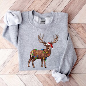 Puede incluir: Una sudadera gris con un reno navide&ntilde;o que lleva un gorro de Pap&aacute; Noel y luces de cadena alrededor de sus cuernos.
