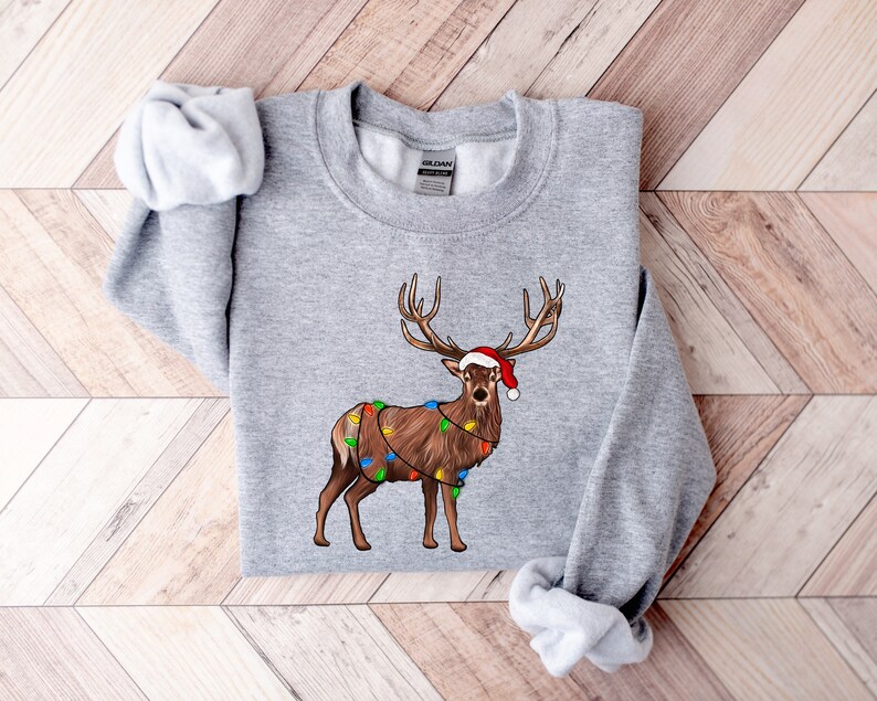 Puede incluir: Una sudadera gris con un reno navide&ntilde;o que lleva un gorro de Pap&aacute; Noel y luces de cadena alrededor de sus cuernos.