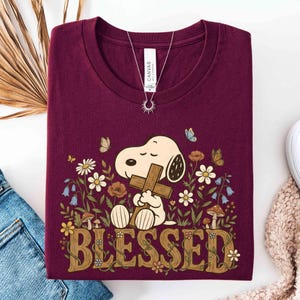 May include: Burgundy t-shirt featuring Snoopy holding a cross, surrounded by flowers and butterflies. The word "BLESSED" is displayed in a decorative font. A silver necklace with a sun pendant is visible.