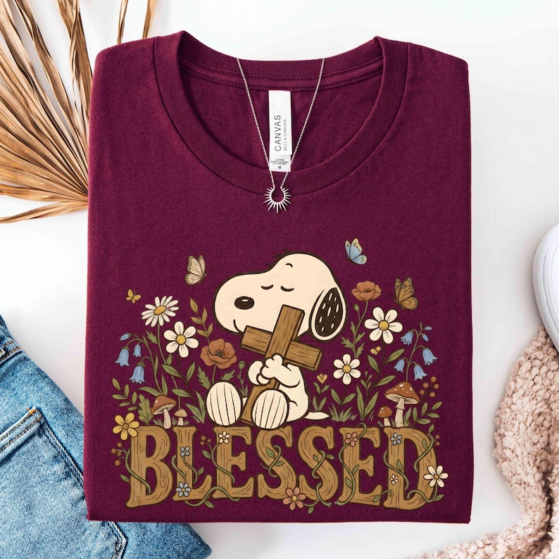 Puede incluir: Camiseta burdeos con Snoopy sosteniendo una cruz, rodeado de flores y mariposas. La palabra "BLESSED" se muestra en una fuente decorativa. Se ve un collar plateado con un colgante de sol.
