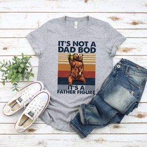 May include: A gray t-shirt with a vintage-style graphic of a bear holding a beer. The text on the shirt reads "It's not a dad bod, it's a father figure."