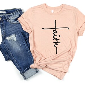 May include: A light pink t-shirt with a black cross and the word "faith" written vertically down the cross.