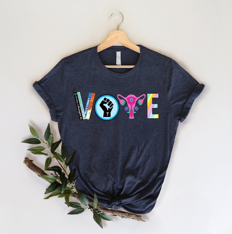 May include: A dark blue t-shirt with the word "VOTE" spelled out in colorful graphics. The "V" is made of books, the "O" is a black fist in a blue circle, the "T" is a pink uterus with flowers, and the "E" is a rainbow.