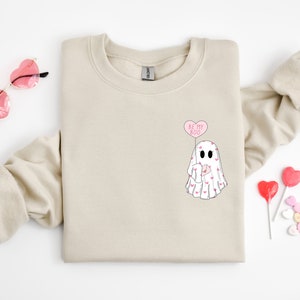 May include: A beige sweatshirt with a ghost graphic and the text "Be My Boo" in a pink heart-shaped balloon. The ghost is holding a pink heart and is covered in pink hearts.