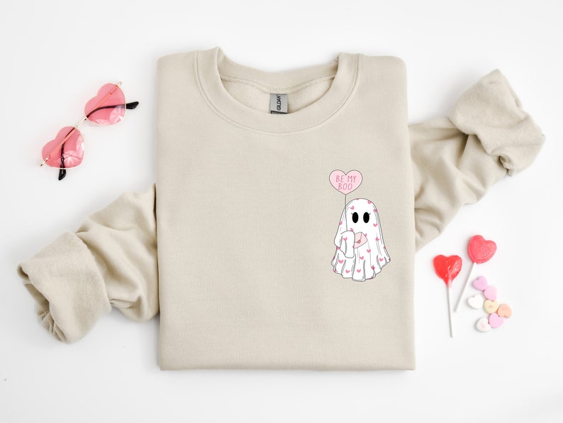 May include: A beige sweatshirt with a ghost graphic and the text "Be My Boo" in a pink heart-shaped balloon. The ghost is holding a pink heart and is covered in pink hearts.