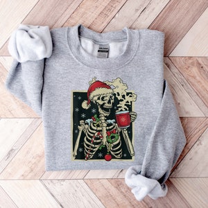 May include: A gray sweatshirt with a Christmas-themed graphic of a skeleton wearing a Santa hat and holding a mug of hot cocoa. The skeleton is decorated with candy canes, holly, and ornaments.