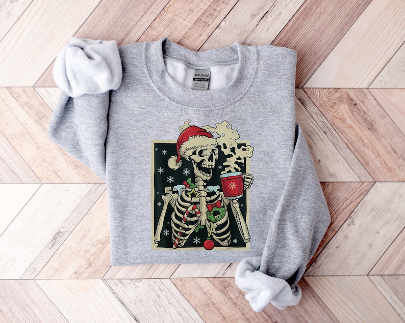 May include: A gray sweatshirt with a Christmas-themed graphic of a skeleton wearing a Santa hat and holding a mug of hot cocoa. The skeleton is decorated with candy canes, holly, and ornaments.