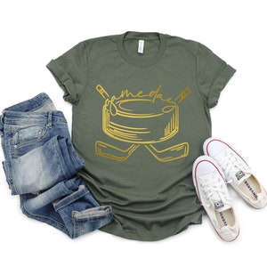 May include: A green t-shirt with a gold graphic of a hockey puck and two hockey sticks. The text "game day" is written above the puck.