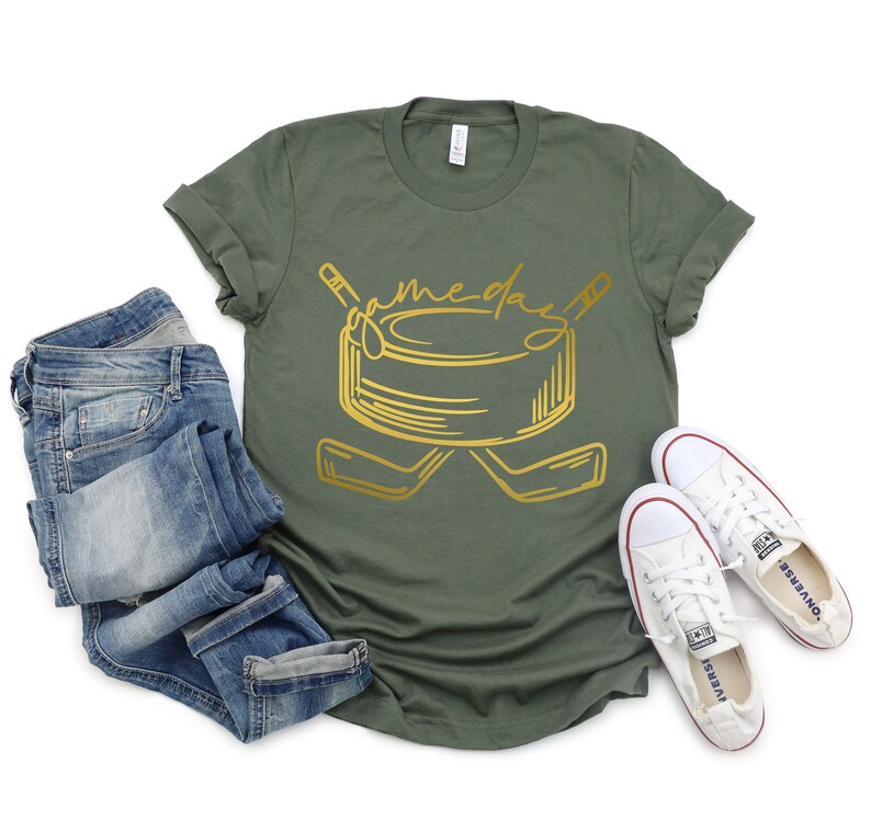 May include: A green t-shirt with a gold graphic of a hockey puck and two hockey sticks. The text "game day" is written above the puck.
