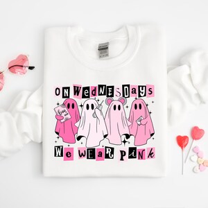 May include: A white crewneck sweatshirt featuring a graphic of four ghosts in various shades of pink and white. The text reads "ON WEDNESDAYS WE WEAR PINK" in black and pink lettering. The ghosts are holding a book, a phone, and a letter.