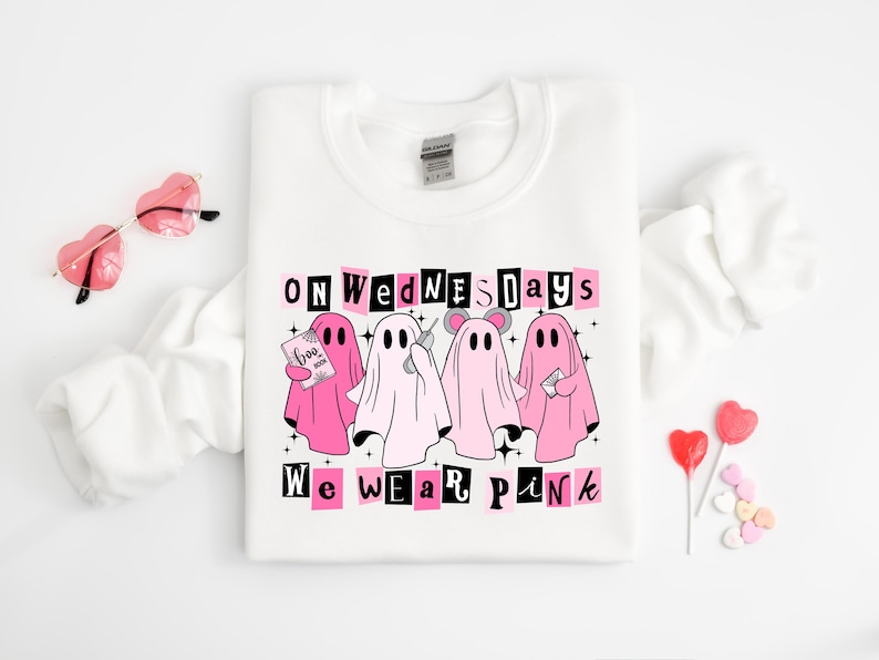 May include: A white crewneck sweatshirt featuring a graphic of four ghosts in various shades of pink and white. The text reads "ON WEDNESDAYS WE WEAR PINK" in black and pink lettering. The ghosts are holding a book, a phone, and a letter.
