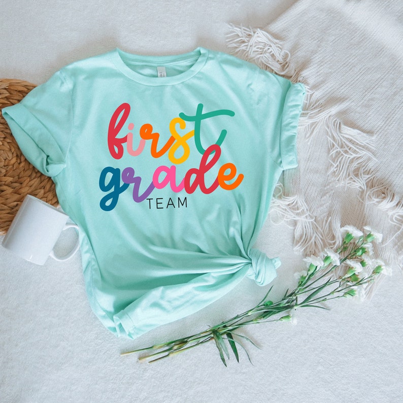 K&ouml;nnte beinhalten: Ein hellblaues T-Shirt mit dem Text "first grade team" in Regenbogenfarben.