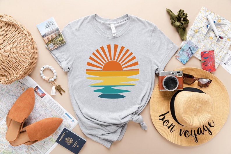 May include: A gray t-shirt with a retro sunset graphic. The graphic features a stylized sun with orange, yellow, and blue colors. The shirt is laid out on a table with other travel-themed items, including a straw hat, a passport, and a map.
