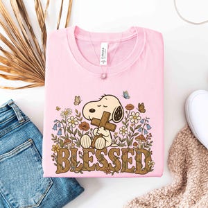Puede incluir: Camiseta rosa claro con un dibujo de Snoopy sosteniendo una cruz, rodeado de flores y mariposas. La palabra "BLESSED" est&aacute; escrita en marr&oacute;n con motivos florales. La camiseta est&aacute; doblada, junto a unos vaqueros azules y zapatos blancos.
