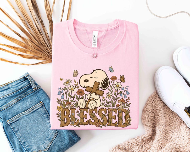 Puede incluir: Camiseta rosa claro con un dibujo de Snoopy sosteniendo una cruz, rodeado de flores y mariposas. La palabra "BLESSED" est&aacute; escrita en marr&oacute;n con motivos florales. La camiseta est&aacute; doblada, junto a unos vaqueros azules y zapatos blancos.