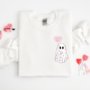 May include: White sweatshirt with a ghost holding a heart-shaped balloon that says "Be My Boo". The ghost is covered in small pink hearts.