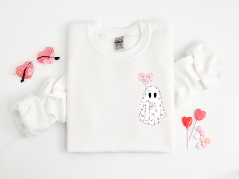 May include: White sweatshirt with a ghost holding a heart-shaped balloon that says "Be My Boo". The ghost is covered in small pink hearts.