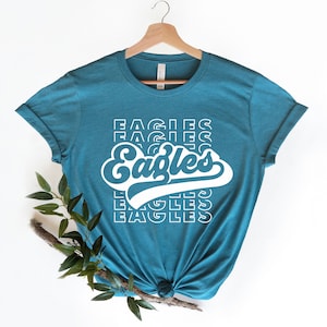 以下が含まれることがあります： ターコイズブルーのTシャツに、レトロなスタイルで「イーグルス」という単語が白で書かれたグラフィックがプリントされています。