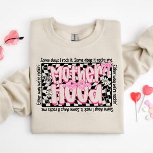 May include: A beige sweatshirt with a black and white checkered graphic that says "Mother Hood" in pink letters. The graphic is surrounded by pink hearts, flowers, and a pink bow. The text around the graphic says "Some days I rock it. Some days it rocks me. Either way we're rockin'. Some days I rock it. Some days it rocks me."