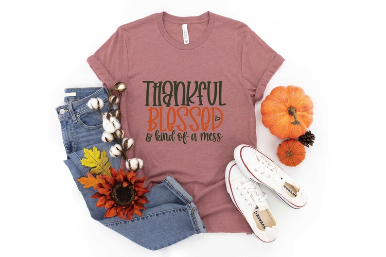 May include: A mauve t-shirt with the text "Thankful Blessed & kind of a mess" in black and orange lettering. The shirt is displayed on a white background with jeans, a sunflower, and pumpkins.
